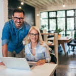 Two smiling business professionals working together on a laptop to build business credit in a modern office.