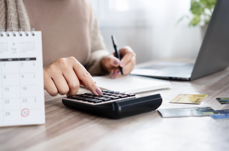 A person using a calculator at a desk with credit cards, a notebook, and a calendar nearby, illustrating concepts related to financial planning and CPN tradelines for sale.